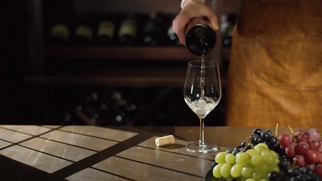 The sommelier is slowly filling the glass with white wine. The lovely composition of the colourful juicy grapes placed on the black plate and the shadows of the window.