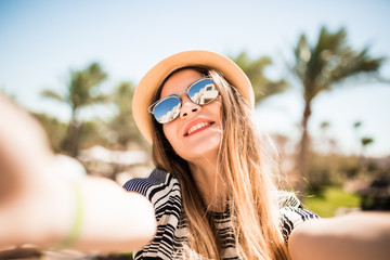 Smile woman in hat and sunglasses taking selfie with mobile phone from hands on summer resort palms background. Summer vocation