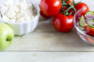 Foods for healthy eating: cottage chesse, tomatoes, apple, salad on light wood background