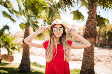 Cheerful lovely young woman in red dress and hat, sunglases walking and talking on mobile phone on summer resort