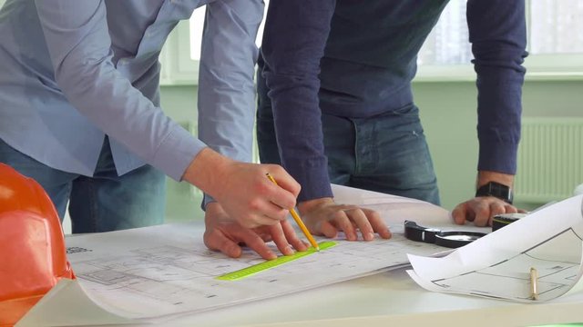 Two young architects making marks on the plans of the building. Close up of male hands using pencil and ruler to draw lines. Construction engineers working with project documentations