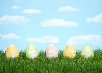 Row of pastel colored Easter eggs swirled design laying on grass with blue background, sky with clouds. Copy space