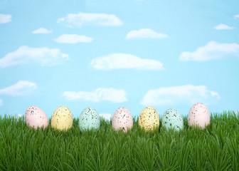 Row of pastel colored Easter eggs splattered with speckles of paint laying on grass with blue background, sky with clouds. Copy space.