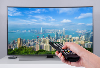 Hand holding TV remote control with a television and city screen in the background