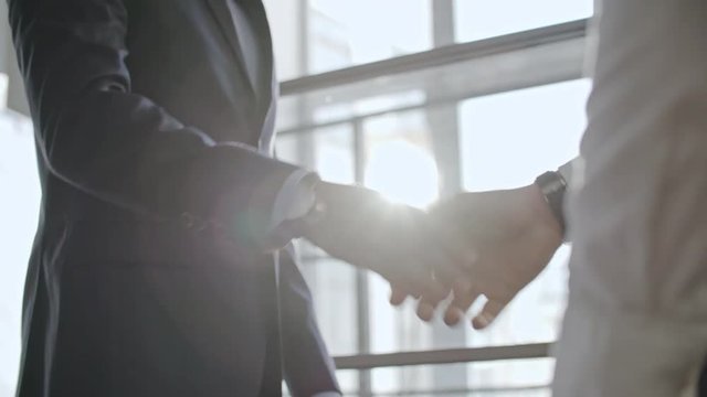 Low angle shot with tilt up of two businessmen in official clothes greeting each other with handshake and discussing work before big window
