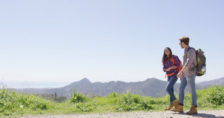 Young laughing couple wearing backpacks and walking down road while hiking in mountains in summer time. 