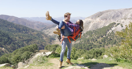 Young loving man holding girlfriend on hands and both laughing with mountainscape on background.