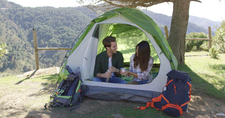 Young laughing couple sitting in tent and drinking tea while taking rest in mountains. 