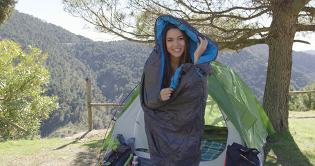 Young laughing female having fun and posing in sleeping bag with tent and mountains on background. 