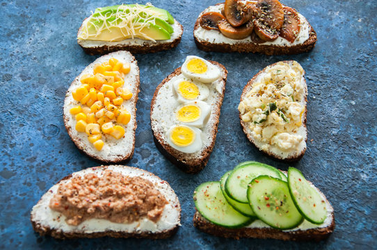 Mix of rye bread bruschettas with various filling. Sweet corn Avocado Mushrooms Quail Eggs Cucumber Omelette Tuna Seeds Cheese sandwiches on blue background. Flat lay.