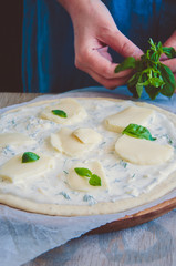Obraz na płótnie Canvas White pizza dough ready to bake on parchment paper over round wooden board. Woman's hand putting the basil leaves on a dough.