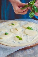 White pizza dough ready to bake on parchment paper over round wooden board. Woman's hand putting the basil leaves on a dough.