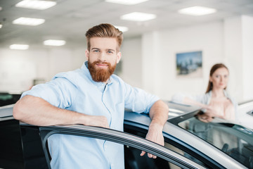 Happy couple standing at car in dealership salon, man looking at camera