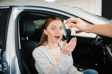 Person giving car key to excited woman sitting in car in dealership salon