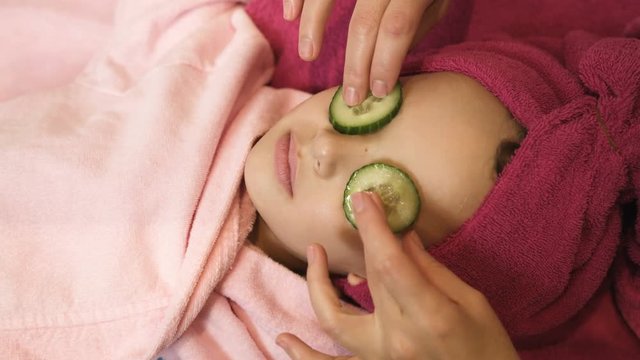 Professional beautician putting cucumber slices on little girls eyes at beauty salon. Spa therapy. Close up view
