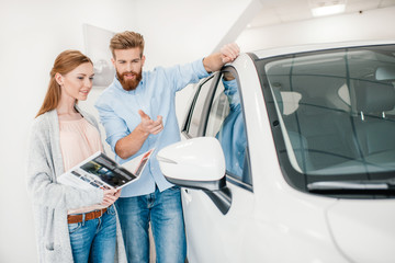 Happy couple holding catalog and choosing car in dealership salon