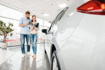 Happy couple holding catalog and choosing car in dealership salon