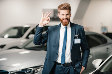 salesman in suit showing ok sign and looking at camera in dealership salon