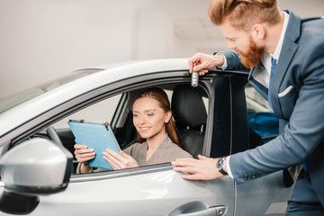 Bearded salesman giving car key to young woman sitting in new car with digital tablet