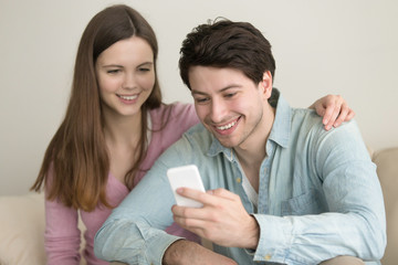 Young happy couple at home using smartphone, making video call, hand on shoulder, online shopping, downloading apps, online banking, messaging, social networking, planning vacation, booking, talking