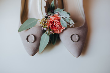 Wedding rings lie on the bride's shoes next to the boutonniere of the groom of red flowers and eucalyptus