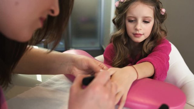 Nail artist doing manicure for pretty little girl with curls in professional salon. Close up view of manicure master applying gel polish