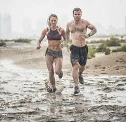 Muscular male and female athlete covered in mud running down a rough terrain with a desert...