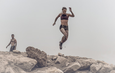 Strong beautiful muscular female athlete covered in mud jumping of a rough rocky ledge capture in mid air.