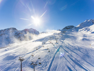 Skiing in the Alps with snow canons throwing fresh powder. Lens flare visible.