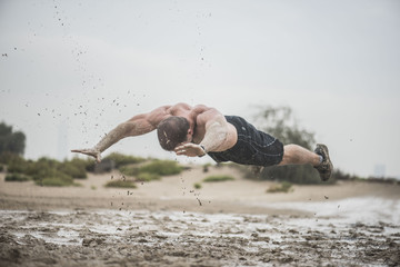 A powerful strong athletic male does a flying pushup in a muddy puddle with a rough terrain with a city in the background