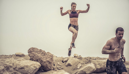 Strong beautiful muscular female athlete covered in mud jumping of a rough rocky ledge capture in...