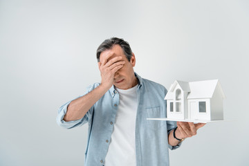 portrait of upset mature man holding house model on grey