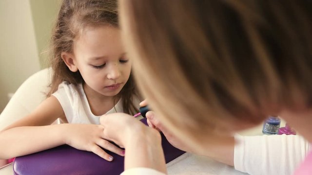 Nail master doing manicure for pretty little girl in professional salon. Close up view of manicure artist applying gel polish
