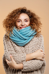 Close up portrait of cheerful smiling beautiful brunette curly girl in knitted sweater and grey neckwarmer over beige background.