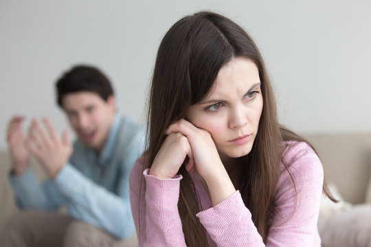 Unhappy couple having a dispute, sitting apart at home. Man shouting at woman and gesturing. Angry upset lady sitting her back to boyfriend and ignoring, not talking to guy. Troubles in relationships