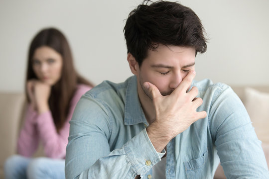 Young guy is very upset and crying, feeling guilty, depressed man in pain, girlfriend sitting in the background indoors, family relationships problems concept, businessman having troubles at work