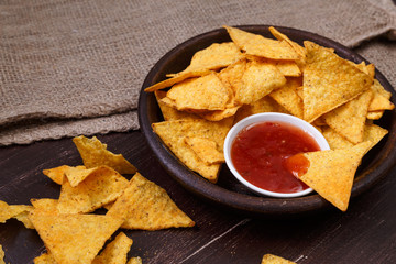 Nachos chips. Delicious salty tortilla with sweet salsa or chilli sauce. Snack on rustic plate. Sackcloth on wooden background.