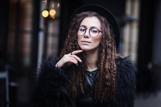 Portrait of a beautiful girl with brown eyes in glasses in a black hat and coat with fur in the cityscape looking in camera. The girl is like Harry Potter