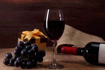 Red wine bottle. Wineglass with dark grapes branch. Gift boxes with bows on rustic wooden background.