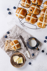 Hot cross buns on baking rack served with butter, fresh blueberries, knife and jug of cream on textile napkin over white texture concrete background. Top view, space. Easter baking.