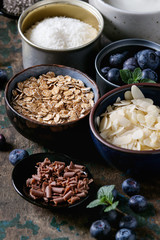 Ingredients for making smoothie for healthy breakfast. Bowls of yogurt, blueberries, granola, almond chia seeds, coconut, milk, chocolate, mint, carambola over dark wooden background. close up