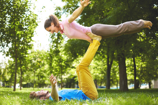 Fit young couple doing acro yoga in park. Man lying on grass and balancing woman in his feet. Happy couple on vacation. Lovers are laughing. Happy guy and girl. Lovers enjoy each other in the city