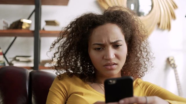 young curly latin woman frown using mobile phone because get bad message and fake new in internet or social network typing messages sitting at home on comfort sofa at home during sunny day