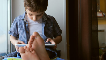 PORTRAIT: Cute little boy sits on a windowsill at home and touches a tablet PC © Cliplab