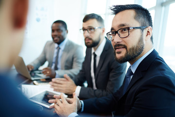 Portrait of middle-aged Asian businessman in eyeglasses sitting at meeting table and listening to his male colleague with attention, profile view