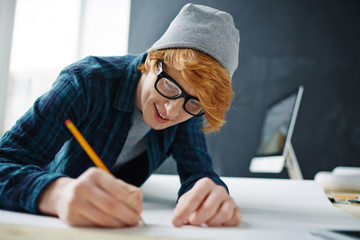 Portrait of young cheerful red haired artist wearing glasses and beanie hat leaning on desk drawing...