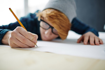 Closeup portrait of young modern red haired man wearing glasses and beanie hat leaning on desk working at blueprints, focus on male hand drawing with pencil on paper