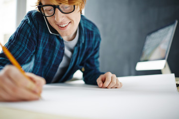 Portrait of young ginger creative student working at blueprints, drawing sketches on paper and talking by phone smiling cheerfully at workplace with modern computer