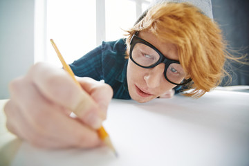 Portrait of young creative red haired man wearing beanie hat and glasses drawing with pencil...
