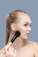 beautiful blond woman holding makeup brush on a gray background. woman applying blusher